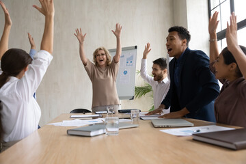 Excited diverse employees team celebrating teamwork success, raising hands and screaming with joy, overjoyed business people rejoicing business achievement, great result in modern boardroom
