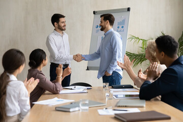 Confident executive businessman wearing glasses shaking successful employee hand, congratulating with job promotion or thanking for work result at corporate meeting, diverse colleagues applauding