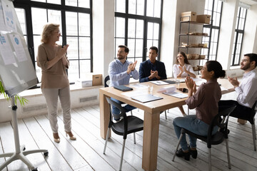 Diverse employees applauding, thanking middle aged mentor for flip chart presentation at briefing,...