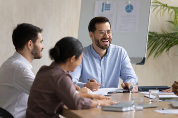 Happy diverse employees with excited team leader having fun at birefing, sitting at table in boardroom, smiling business partners colleagues discussing project, brainstorming, enjoying teamwork