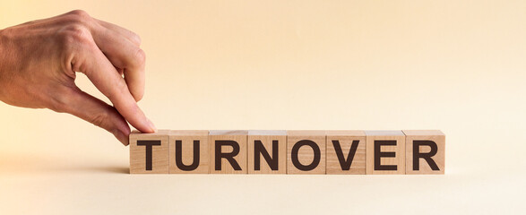 The hand puts a wooden cube with the letter T from the word TURNOVER. The word is written on wooden cubes standing on the yellow surface of the table. Concept.