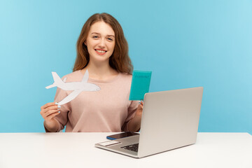 Friendly woman, travel agent holding passport and paper airplane, looking at camera with toothy smile, advertising hot tours with airline transportation. indoor studio shot isolated on blue background