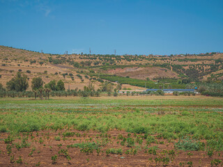 Fototapeta na wymiar Landscape of country area of northern Israel. In the background, there are houses and electricity poles on the mountains and warehouses with solar panels near green agricultural fields in the valley.