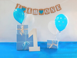 One year birthday decorations set. Decoration for a boy holiday party. Bunch of blue & white balloons, gift boxes, large white kappa board number one shape, and sign with the inscription I am one"."