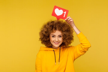Internet blogging. Portrait of happy smiling curly-haired young woman in urban style hoodie holding heart like icon over head, social media button. indoor studio shot isolated on yellow background