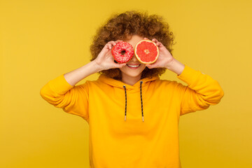 Sugary dessert vs fruits. Portrait of happy curly-haired woman in urban style hoodie covering eyes with fresh grapefruit and sweet doughnut, junk food. indoor studio shot isolated on yellow background