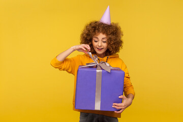 Portrait of happy curly-haired woman unpacking huge gift box with expression full of great expectation, enjoying unwrapping enormous birthday present. indoor studio shot isolated on yellow background