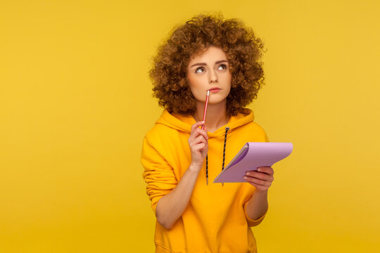 Portrait of pensive curly-haired hipster woman in urban style hoodie thinking over smart idea, holding pencil and notebook to write plans, to-do list. indoor studio shot isolated on yellow background