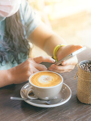 Hot cappuccino coffee for quarantined woman to prevent the spread of the Coronavirus(Covid 19). Woman wearing face mask relaxing with coffee and mobile phones. New normal. Selective focus on coffee.