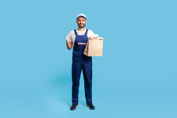Full length positive courier in blue uniform holding groceries bag and showing thumbs up, like gesture, carrying parcel with goods ordered online. Professional delivery service. studio shot isolated