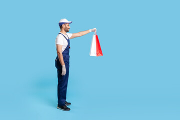 Full length profile of professional courier in uniform giving shopping bags, delivering parcel with goods ordered online at fashion store. indoor studio shot isolated on blue background, copy space