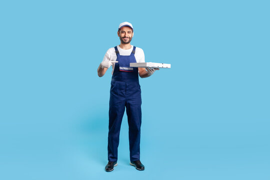 Full length positive professional courier in workwear and protective gloves pointing at pizza box, smiling friendly to camera. Fast food delivery service. studio shot isolated on blue background