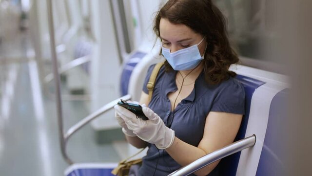 Young woman in medical face mask and protective gloves absorbed in her smartphone while traveling in subway car. 