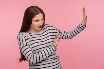 Side view of scared woman in striped sweatshirt raising hands from sudden fear, showing stop, no, refusal gesture, looking frightened, denying problems. indoor studio shot isolated on pink background