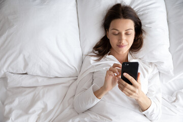 Top view of smiling young woman awaken in white cozy bed using modern smartphone gadget, happy millennial female wake up lie in comfortable bedroom browsing Internet on cellphone in the morning - Powered by Adobe