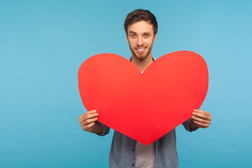 Obraz na płótnie Canvas Portrait of handsome brunette man with bristle holding red paper heart and smiling to camera with alluring flirtatious expression, demonstrating love. indoor studio shot isolated on blue background