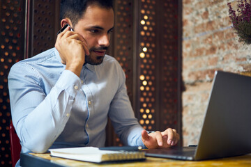 Handsome skilled copywriter discussing ideas for working task having phone talk sitting near laptop...