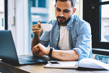 Young caucasian male it developer looking at wristwatch during coffee break sitting in coworking...