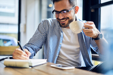 Cheerful male journalist in trendy glasses happy to finishing work on book review rereading written in notebook text before calling to editor to present publication drinking coffee for lunch in cafe