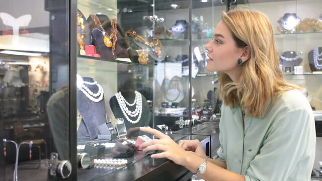 Portrait Cheerful girl choosing natural stone necklace in jewerely shopof young woman looking at showcase with natural stone jewelry . High quality FullHD footage