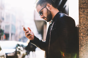Thoughtful man in trendy wear holding smartphone watching online video from social networks standing on street,hipster guy reading notification on telephone making banking spending time outdoors