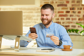 A man finds an online store on a smartphone holding a credit card in his hand in front of the computer at home. A guy doing an online payment on the internet on a laptop in his apartment.