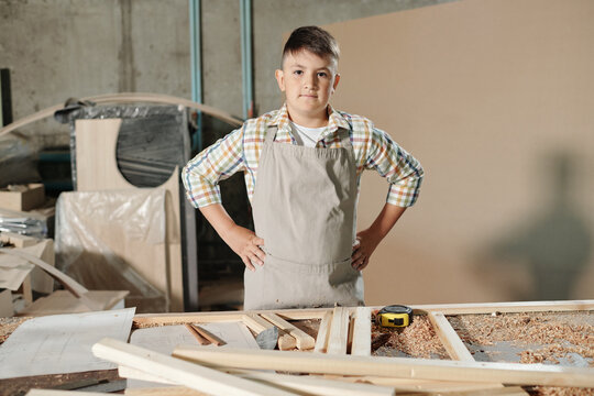 Portrait of serious confident teenage boy in apron holding hands on hips at desk with wooden pieces and shavings in carpentry shop
