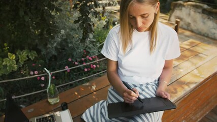 Young lady sitting on bench of city park and drawing on a black screen of digital tablet by stylus, using laptop. Freelance, slow motion, copy space