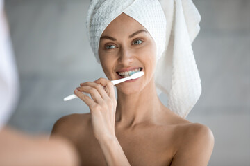 Smiling millennial woman in bath towel after shower look in mirror brush white healthy teeth using...