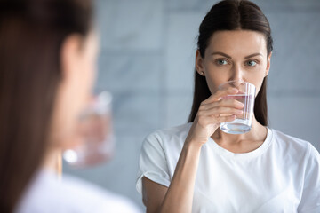 Young woman look in mirror drink clear still mineral water from glass, millennial female enjoy...