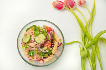 One bowl of salmon pasta salad and a few red tulips flowers in withe background.