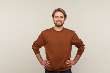 Portrait of positive confident handsome man with neat hair and beard wearing sweatshirt standing, holding hands on hips and looking at camera with toothy smile. studio shot isolated on gray background