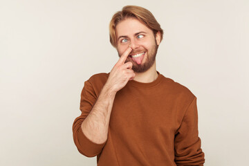 Portrait of dumb brainless comical man with beard in sweatshirt picking nose, looking cross eyed with humorous stupid expression, drilling booger mucus. indoor studio shot isolated on gray background