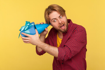 Delighted happy young man using party cone to listen what's inside gift box, curious about present bonus, in anticipation of best birthday surprise. indoor studio shot isolated on yellow background