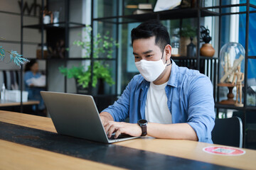 Young Asian Man in blue shirt with face mask working with laptop in coffee shop cafe smile and happy face