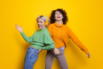 Two happy sisters with curly hair laughing while wearing headphones on a yellow studio wall