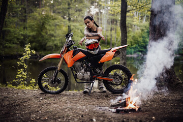 Fototapeta na wymiar Sensual tattooed racer girl wearing motocross outfit with semi naked torso leaning on her bike and looking on camera in the woods