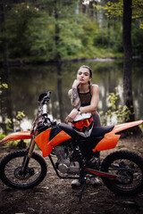 Naklejka na ściany i meble Sensual tattooed racer girl wearing motocross outfit with semi naked torso leaning on her bike and looking on camera in the woods