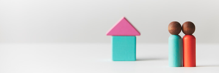 Wooden figurines concept. Two wooden figures people standing in front of a tiny block house, black skin colour, young couple. Realty market. Banner