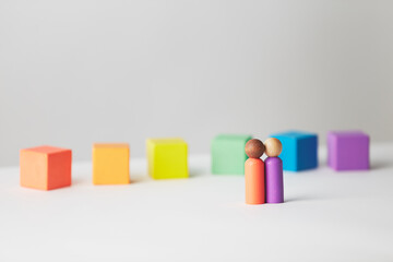 Wooden figures concept. Couple standing in front of wooden blocks in colours of the gay pride rainbow flag. Couple of mixed skin colour. 