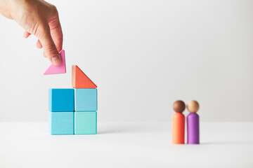Wooden figurines concept. Two wooden figures people standing in front of a large multi generations block house, mixed race skin colour, four adults. Realty market. Human hand finishing the roof.