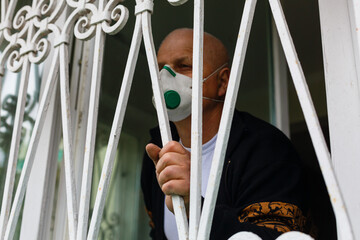 old elderly man in protective face mask against virus looks out through window stay at home for health safety