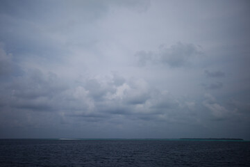 Fototapeta na wymiar Cloudy day in Maldivian ocean with blue grey sky and blue grey water and white send island on the horizont