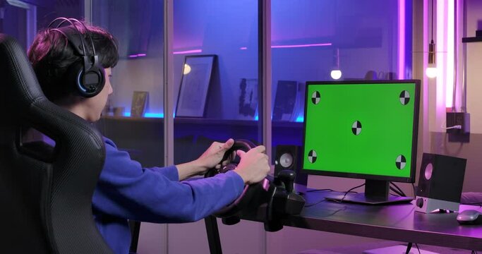 Back view shot of the excited young asian pro cybersport male gamer in headset sitting in the racing seat simulator cockpit with steering wheel and playing with green chroma key screen computer.
