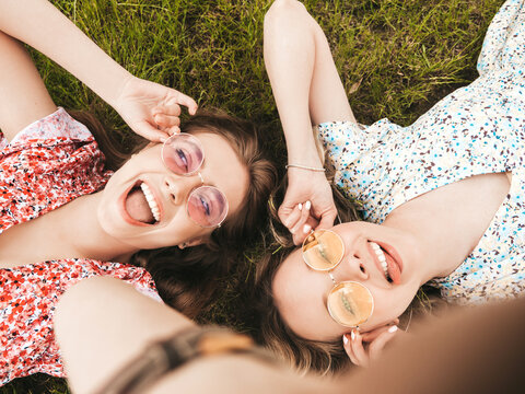 Two young beautiful smiling hipster girls in trendy summer sundress.Sexy carefree women lying on the green grass in sunglasses.Positive models having fun.Top view.Taking selfie photos on smartphone