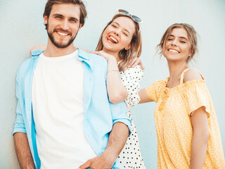 Group of young three stylish friends posing in the street. Fashion man and two cute girls dressed in casual summer clothes. Smiling models having fun near wall.Cheerful women and guy outdoors