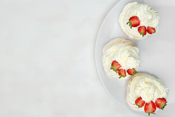 Top view of meringue cakes with cream and halves of strawberries on the plate on a white wooden background with copy space. Delicious dessert