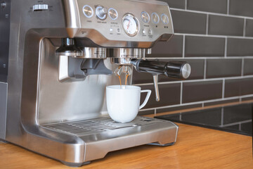 Espresso is poured into a cup while making espresso in a coffee machine (maker) in the kitchen with a black wall and wooden worktop at home.