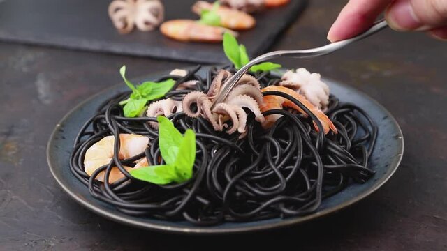 male hand prick an octopus with fork from black cuttlefish ink pasta with shrimps or prawns and basil on a black concrete background. Side view, close up.