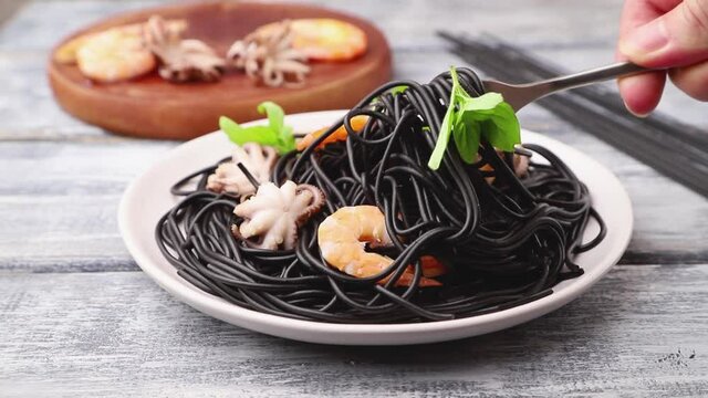 black cuttlefish ink pasta with shrimps or prawns, small octopuses and basil stir with a fork on gray wooden background. Side view, close up.